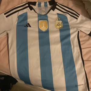 Argentina fotbollströja Adidas  - Säljer en officiell Argentina fotbollströja från Adidas i storlek xs. Tröjan har klassiska blå och vita vertikala ränder, svarta detaljer och broderade märken med AFA-logga och FIFA World Champions 2022-emblem. Tillverkad i lätt och ventilerande material.