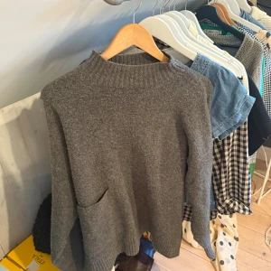 Grå stickad tröja från Zara - Mysig grå stickad tröja från Zara med hög ribbad hals och en praktisk ficka framtill. Tröjan har långärmade ärmar och en loose passform som gör den extra skön. Perfekt för kyliga dagar och enkel att matcha med jeans eller kjol.