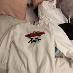 Vit Pontiac sweatshirt från H&M Divided - Vit sweatshirt från H&M Divided med tryck av en röd Pontiac-bil och svart text på bröstet. Tröjan har rund halsringning och långa ärmar. Mjuk insida och klassisk passform, perfekt för dig som gillar retrobilar och streetstyle.