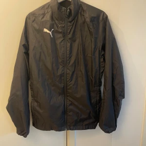 Svart Puma vind- & regnjacka – sportig & praktisk! - Svart jacka från Puma i lätt polyester, perfekt för regniga och blåsiga dagar. Dragkedja framtill, hög krage och huva som enkelt kan vikas in. Sportig och tidlös design med logga på bröstet – både praktisk och snygg i alla väder!