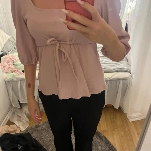 Rosa blus med knyt från 157 - Supersöt ljusrosa blus från 157 med trekvartsärmar och knyt i midjan. Blusen har en fyrkantig ringning och lätt vågig nederkant. Materialet är mjukt och har en lätt struktur som ger en chill vibe. Perfekt för dig som gillar feminina detaljer och en avslappnad look.