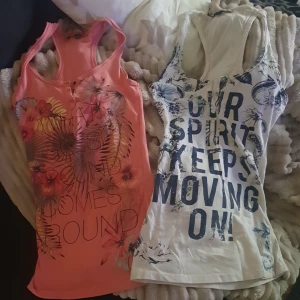 2 Bershka linnetoppar med tryck S - Två snygga linnetoppar från Bershka. Ena toppen är korallrosa (S) med blommigt tryck och knappar framtill, den andra är vit (XS)med blå text och blommönster samt knappar. Båda är ärmlösa och har racerback-modell.