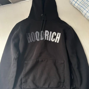Svart Hoodie  Hoodrich  - Svart hoodie från Hoodrich med stor broderad logga i silvergrått på bröstet. Tröjan har huva med snörning, magficka och långa ärmar. Perfekt för dig som gillar streetwear och vill ha en snygg och bekväm look.