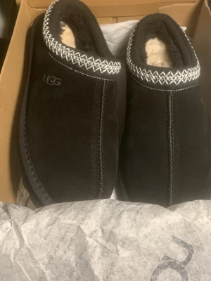 Svarta UGG mockatofflor med mönster - Svarta tofflor från UGG i mjuk mocka med fluffigt foder och dekorativt vitt zigzag-mönster runt öppningen. Perfekta för att hålla fötterna varma och sköna. Klassisk UGG-logga på sidan och robust sula för extra komfort.