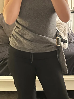 Grå ärmlös omlottopp med bälte - Säljer en stilren grå ärmlös topp med omlottdesign och ett brett bälte med guldfärgat spänne i midjan. Toppen har en mjuk, stretchig känsla och sitter snyggt figurnära. Perfekt till jeans eller kjol för en trendig look.