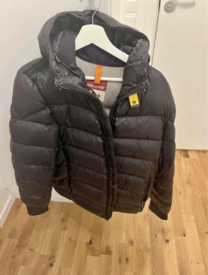 Svart dunjacka från Parajumpers - Säljer en svart dunjacka från Parajumpers med huva och gul detalj vid halsen. Jackan har broderad logga på ärmen, dragkedja framtill och två sidofickor. Insidan är ljusgrå med meshficka och tydlig Parajumpers-label. Perfekt för kalla dagar.