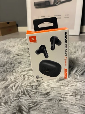 JBL Wave Beam 2 - JBL Wave Beam 2 trådlösa hörlurar med aktiv brusreducering, djup bas och upp till 40 timmars batteritid. IP54-klassade, levereras med laddningskabel och originalförpackning. Produkten var svår att komma in i så förpackningen är förstörd på toppen.