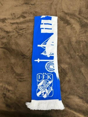 IFK Göteborg blåvit halsduk - Säljer en blå och vit halsduk från IFK Göteborg med klubbens logga och Göteborgs siluett invävd. Halsduken har vita fransar i ändarna och är perfekt för att visa ditt stöd på matchen. Klassisk supporterstil för dig som älskar fotboll.