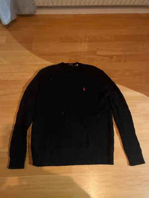 Svart stickad tröja från Polo Ralph Lauren - Svart kabelstickad tröja från Polo Ralph Lauren med klassisk rund hals och långärmad passform. Tröjan har en röd broderad logga på bröstet och ribbade muddar vid ärmslut och nederkant. Perfekt för dig som gillar stilrena och tidlösa plagg. Är i s men är lite större i sin storlek
