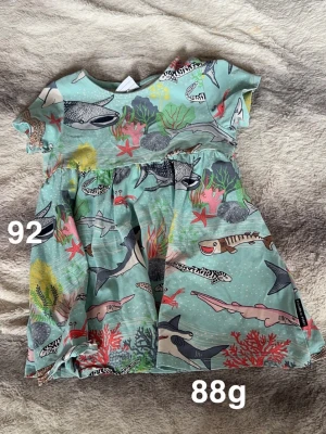 Kort klänning med hajmönster - Supersöt kort klänning från Polarn o pyret i ljusblått med färgglatt undervattensmönster med hajar, koraller och sjöstjärnor. Perfekt för dig som gillar lekfulla prints och bekväma plagg.🌸 Fint använt skick. Storlek 92