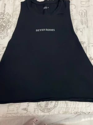 Svart Better Bodies linnetopp - Säljer en svart linnetopp från Better Bodies med logga tryckt framtill. Toppen är ärmlös och har en sportig, tight passform som passar perfekt till träning. Materialet känns lätt och stretchigt, vilket gör den bekväm att bära under intensiva pass.