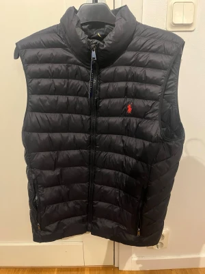 Svart dunväst från Polo Ralph Lauren - Säljer en svart dunväst från Polo Ralph Lauren med quiltad design och dragkedja framtill. Västen har ståkrage, två fickor med dragkedja och den klassiska röda loggan broderad på bröstet. Perfekt att slänga över en hoodie eller tröja för extra värme. Den är i  bra skick och köpt från Polo Ralph laurens egna hemsida 