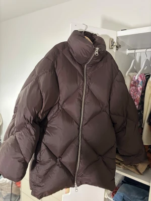 Brun oversized pufferjacka - Other stories  - Säljer en mörkbrun pufferjacka från: Other stories.  Jackan har hög krage, stora quiltade partier och dragkedja framtill. Slutsåld överallt!  nypris 3200kr! Säljes nu för 1400kr. Storlek: XS. Passar både XS och S. 