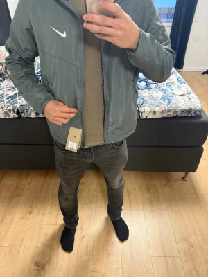 Grå Nike vindjacka med huva - Säljer en grå Nike vindjacka med huva och dragkedja framtill. Jackan har en clean look med vit Nike-logga på bröstet och praktiska sidofickor. Perfekt för löprundan eller när du vill ha en lätt jacka till vardags. Tillverkad i polyester.