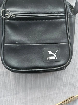 Svart axelväska från Puma - Säljer en svart axelväska från Puma i syntetmaterial med klassisk logga framtill. Väskan har en dragkedja upptill, ytterfack med dragkedja och ett grönt mönstrat innerfoder med Puma-tryck. Perfekt storlek för det viktigaste och enkel att bära över axeln.