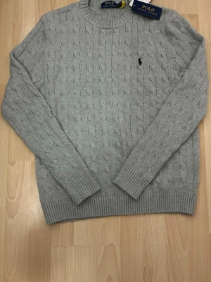 Grå stickad Polo Ralph Lauren tröja - Säljer en grå kabelstickad tröja från Polo Ralph Lauren med klassisk rund hals och svart broderad logga på bröstet. Tröjan har ribbade muddar vid ärmslut och nederkant samt lång ärm. Perfekt för dig som gillar stilren och tidlös design.