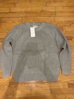 Grå stickad tröja Maison Margiela - Säljer en ribbstickad grå tröja från Maison Margiela. Tröjan har klassisk rund halsringning, långa ärmar och den ikoniska vita sömmen i nacken. Perfekt för dig som gillar minimalistisk och stilren design.