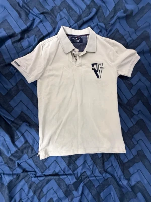 Vit pikétröja från iSEE D.N.M - Klassisk vit pikétröja från iSEE D.N.M med korta ärmar och krage. Tröjan har en broderad logga med texten 'Vintage' och 'TM REG' på bröstet. Tillverkad i bomull för en skön och luftig känsla. Perfekt till jeans eller shorts för en clean look.