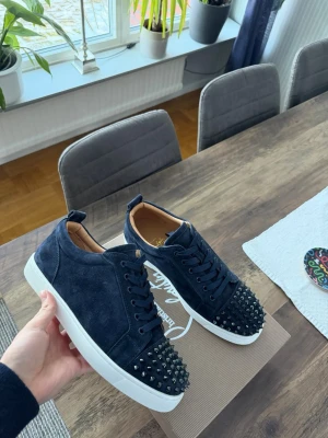 Mörkblå mocka sneakers med nitar - Säljer ett par mörkblå sneakers från Christian Louboutin i mjuk mocka. Skorna har vita sulor, snörning och coola svarta nitar över hela tån för en edgy vibe. Insidan är beige och märkt med guldtext. Perfekta för dig som vill sticka ut.