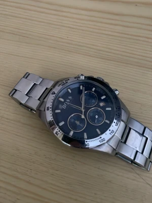 Snygg silverfärgad Hugo BOSS klocka - Stilren klocka från BOSS med silverfärgad länk i metall och mörkblå urtavla. Klockan har kronograf-funktion, datumvisning och tydliga indexmarkeringar. Perfekt accessoar för dig som vill ha en klassisk men modern look. Köpte klockan för 4590 kr för några år sen men jag har tappat den och glaset gick sönder så jag har bytt glaset och rengjort ramen. Klockan fungerar inte så det kostar cirka 1500 och fixa den till en helt ny. Säljer billigt med tanke på att det är en Hugo boss klocka. 