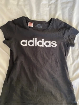Svart Adidas t-shirt med logga - Säljer en svart t-shirt från Adidas med stor vit logga över bröstet. Klassisk rund halsringning och korta ärmar. Tillverkad i mjuk bomull som är skön att ha på sig. Perfekt för dig som gillar sportiga och stilrena plagg.