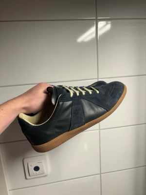 Maison Margiela gats - Säljer ett par snygga Maison Margiela sneakers i mörkblå mocka och skinn med beige snörning och klassisk gummisula. Skorna har en retro siluett med rund tå och kontrasterande materialmix som ger en clean och stilren vibe.