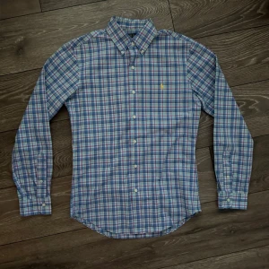 Rutig skjorta från Polo Ralph Lauren - Säljer en slim fit rutig skjorta från Polo Ralph Lauren i blå, vit och röd färg. Skjortan har lång ärm, klassisk krage och knappar framtill. Ikoniska broderade loggan på bröstet. Tillverkad i mjuk bomull för skön känsla.