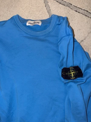 Blå sweatshirt från Stone Island Äkta Storlek M - Säljer en blå sweatshirt från Stone Island med den klassiska patchen på ärmen. Tröjan har rund halsringning och är tillverkad i mjuk bomull. Perfekt för dig som vill ha en clean och stilren look med streetwear-vibe.