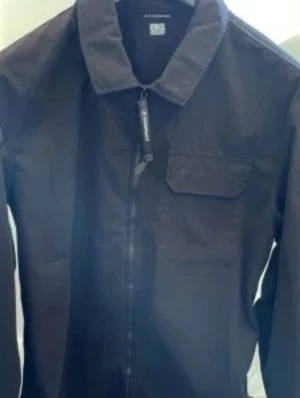 CP Company overshirt - Säljer en svart overshirt från C.P. Company med klassisk krage, bröstficka och ikonisk linsdetalj på ärmen. Jackan har dragkedja framtill och är tillverkad i ett slitstarkt bomullsmaterial. Perfekt för lager på lager och stilren streetwear.