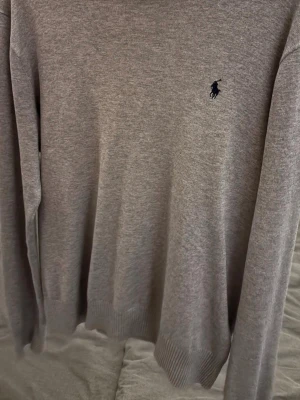 Grå stickad tröja från Ralph Lauren - Säljer en klassisk grå stickad tröja från Ralph Lauren med den ikoniska loggan broderad på bröstet. Tröjan har lång ärm, rund halsringning och ribbade muddar vid ärmslut och nederkant. Perfekt till lager på lager under hösten och vintern.storlek 14-16 motsvarar xs/s skriv vid funderingar. Pris kan diskuteras.