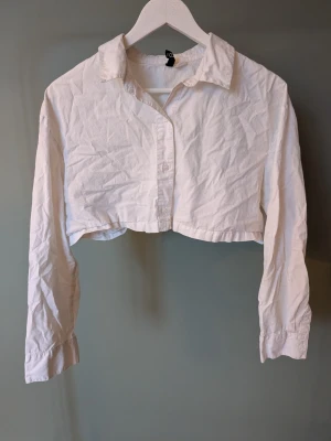 Vit croppad blus från H&M - Säljer en vit croppad blus från H&M i storlek M. Blusen har klassisk krage, knappar framtill och långa ärmar. Tillverkad i mjuk bomull och har en loose passform som ger en chill vibe. Perfekt att styla med höga jeans eller kjol.