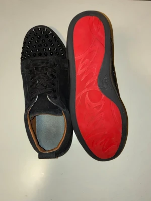 Svarta sneakers med nitar inspirerade av  Louboutin - Säljer ett par svarta sneakers som är inspirerade av Christian Louboutin i mocka med ikoniska röda sulor. Skorna har rund tå, snörning och coola svarta nitar på tån. Insidan är fodrad i beige skinn och sulan har Louboutin-loggan ingraverad. Bra om man vill ha ett bra och snygga skor som inte kostar så mycket (inspirerade av Christian Louboutin) 