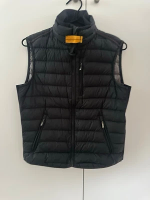 Svart dunväst från Parajumpers - Säljer en svart dunväst från Parajumpers med klassisk quiltad design och hög krage. Västen har dragkedja framtill, tre fickor med dragkedja och den ikoniska orangea detaljen vid halsen. Perfekt för lager-på-lager och riktigt snygg till höst och vinter.!pris kan diskuteras!