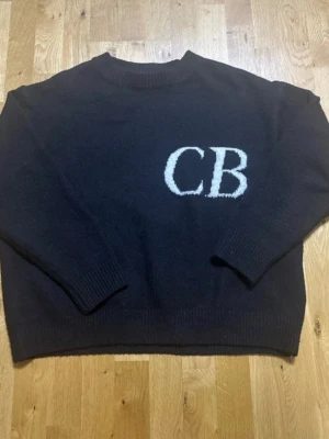 Cole Buxton knit wear  - Säljer en svart stickad tröja från Cole Buxton med stora vita bokstäver 'CB på bröstet. Tröjan har rund hals, ribbade muddar och en loose passform som ger en avslappnad vibe. Perfekt för dig som gillar streetwear.