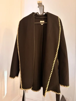 Svart omlottkappa från Kappahl - Svart omlottkappa från Kappahl med beige/vit sicksack-detaljer längs kanterna och bälte i midjan.