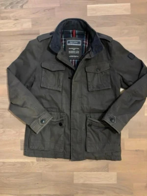 Grön field jacket från Marc O'Polo - Snygg mörkgrön parkajacka från Marc O'Polo med rutigt innerfoder. Jackan har fyra stora fickor framtill, axelklaffar och stängs med dragkedja och knappar. Perfekt för höst och vinter, tillverkad i slitstark bomull och polyester.