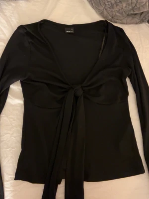 Svart omlottopp med knyt från Glamorous - Säljer en svart omlottopp från Glamorous med långa ärmar och snygg knytning framtill. Toppen har en djup v-ringning och är figurnära i modellen. Perfekt att styla med jeans eller kjol för en trendig look.