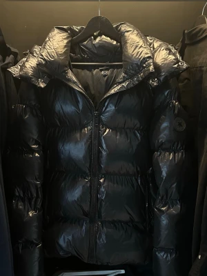 Svart pufferjacka  - Säljer en svart, glansig pufferjacka från Everest med stor huva och dragkedja framtill. Jackan har en klassisk pufferlook med breda kanaler och en patch på ärmen. Perfekt för kalla dagar när du vill vara både varm och stilren. Storlek S passar M
