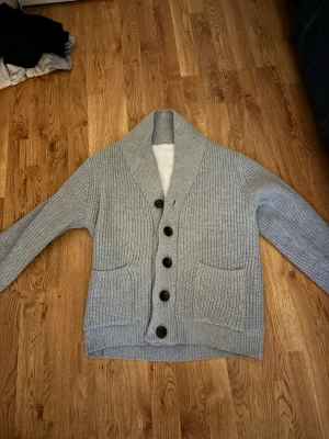 Grå stickad kofta med fuskpälsfoder - Mysig grå stickad kofta med stora svarta knappar och två fickor framtill. Insidan är fodrad med fluffigt vitt fuskpälsfoder som gör den extra varm. Perfekt för kyliga dagar och har en avslappnad, lite oversized känsla. Två små revor som man ser på bild 3 och 4 går och sy ihop. Syns bara på insidan. Storlek m passar mer som s