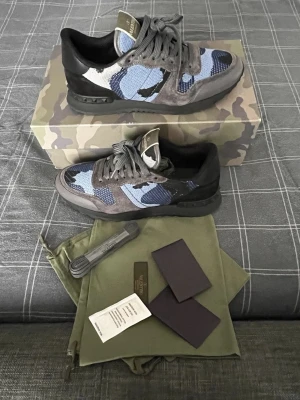 Valentino Garavani Rockrunner sneakers - Säljer ett par Valentino Garavani Rockrunner sneakers i blått och grått med camouflage-mönster. Skorna har detaljer i mocka och mesh, svarta partier och coola nitar på hälen. Klassisk snörning och rund tå. Kommer med originalkartong, extra skosnören och dustbags.