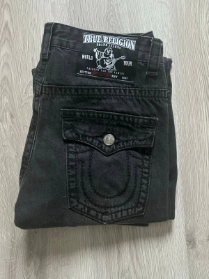 Svarta True Religion jeans, storlek 32 - Säljer ett par svarta jeans från True Religion med ikoniska bakfickor och silverfärgade knappar. Klassisk rak passform och tillverkade i USA. Snygga detaljer på fickorna och cool label bak i midjan. Perfekta för dig som gillar streetwear och vill sticka ut.