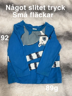 Blå långärmad t-shirt Polarn O. Pyret - Blå långärmad t-shirt från Polarn O. Pyret med ett coolt tryck av en figur. Skön passform, perfekt för lek och vardag. Klassisk rund hals och tryckdetaljer framtill.🌸 Något slitet tryck. Små fläckar. Storlek 92