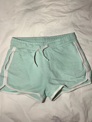 Mintgröna shorts med vit kant - Säljer ett par mintgröna shorts från Peak Performance med vit kant och snörning i midjan. De har en loose passform och är gjorda i mjuk bomull, perfekta för chill dagar eller träning. Klassisk sportig look med rundade ben och elastisk midja! Passar superbra till sommaren, eller bara till en myskväll hemma i soffan!