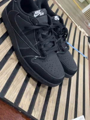 Nike Air Jordan 1 Low Travis Scott svart - Nike Air Jordan 1 Low Travis Scott i svart mocka med vita kontrastsömmar och broderade detaljer på hälen. Skorna har klassisk snörning, rund tå och platt sula. Innersulan har Cactus Jack- och Jumpman-logga. Perfekt för dig som vill sticka ut med en ikonisk sneaker.
