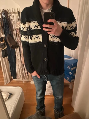 Svart kofta med mönster från George - Mysig svart stickad kofta från George med vita mönster och djurmotiv över bröstet och armarna. Koftan har stora knappar framtill, bred krage och långa ärmar. Perfekt för kalla dagar när du vill vara både varm och snygg.