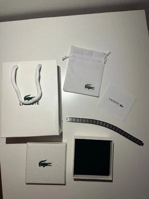 Lacoste silverfärgad armbandslänk - Säljer ett stilrent armband från Lacoste i silverfärgad metall med klassiska krokodilloggan ingraverad på länken. Armbandet har ett mönstrat rutigt motiv på varje länk och kommer med original ask, påse och instruktioner. Perfekt accessoar för dig som gillar snygga detaljer.