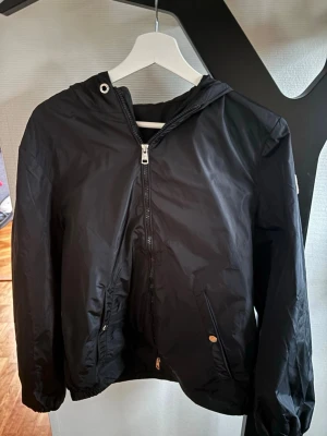 Svart vindjacka från Moncler - OBS: jackan passar mellan S och Xs. Svart windbreaker från Moncler med huva och dragkedja framtill. Jackan har vita och röda ränder på huvan, två fickor med knapp och elastiska muddar vid ärmslut och nederkant. Materialet är lätt och syntetiskt 