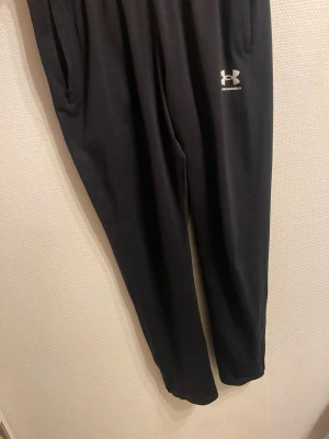 Svarta mjukisbyxor från Under Armour - Svarta mjukisbyxor från Under Armour med elastisk midja och diskret logga på vänster lår. Tillverkade i mjukt syntetmaterial som känns skönt mot huden. Perfekta för chill eller träning. Klassisk rak passform och fickor på sidorna.