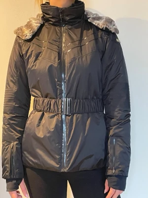 Svart vinterjacka från Colmar med fuskpäls - Snygg svart vinterjacka från Colmar med markerad midja och bälte. Jackan har dragkedja framtill, långa ärmar med dragkedjedetaljer och en stor huva fodrad med mjuk gråmelerad fuskpäls. Perfekt för kalla dagar och riktigt stilren look. Snyggast i skidbacken! 