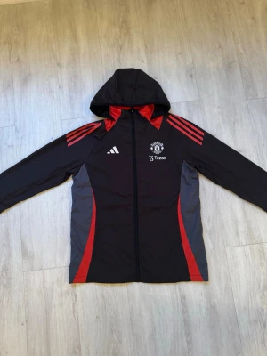 Manchester United vindjacka Adidas - Snygg svart och röd Manchester United windbreaker från Adidas med huva och hel dragkedja. Jackan har röda detaljer och tre ränder på axlarna, samt klubbmärke och Tezos-logga på bröstet. Perfekt för dig som älskar fotboll och vill ha en sportig look. 